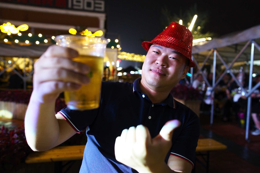 A guy in a sequined hat holds up a beer to the camera and smiles