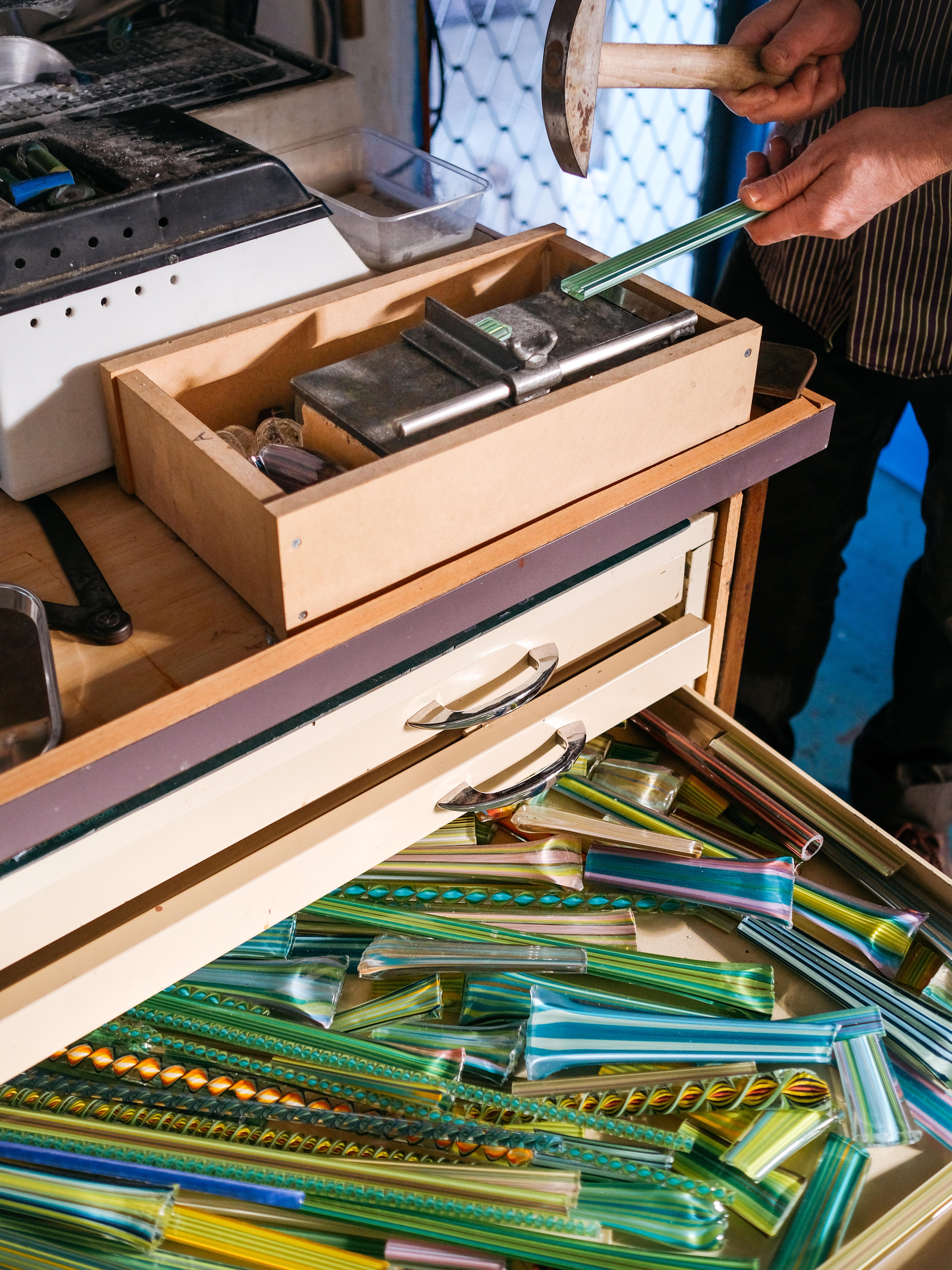 Close up of artist Tom Moore's hands holding a chisel and breaking a piece of glass, above a colourful drawer of glass pieces