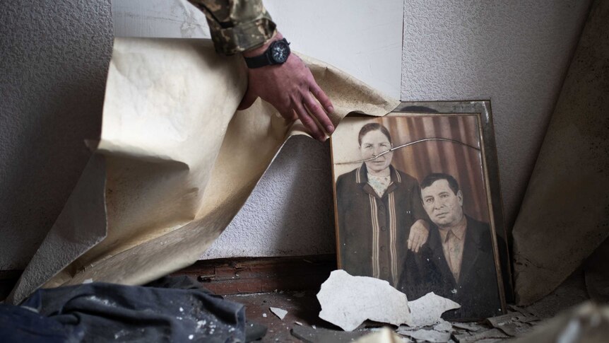 A soldier's hand reveals an old photograph of a man and a woman beside some peeling wallpaper.