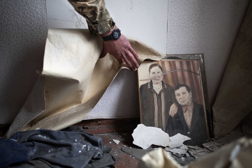 A soldier's hand reveals an old photograph of a man and a woman beside some peeling wallpaper.