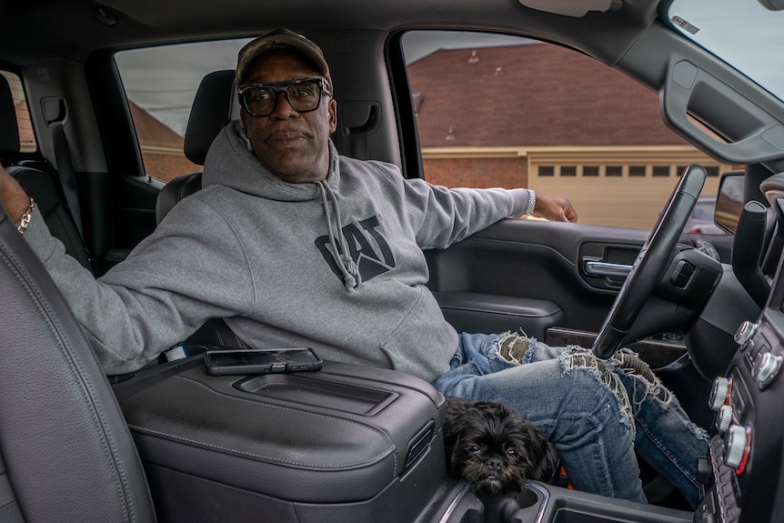 A Black man wearing a grey hoodie and black glasses sits in his car talking.