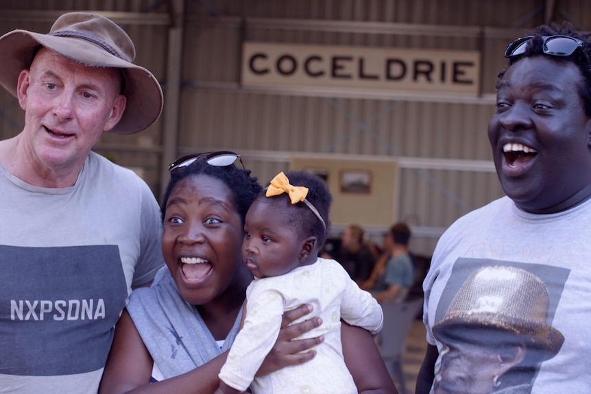 Two men, one woman and a baby all laughing, outdoors in a barn. 