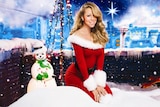 Blonde haired woman wearing a Mrs. Clause dress kneeling in fluffy snow, next to a snowman with a green scarf and white hat.