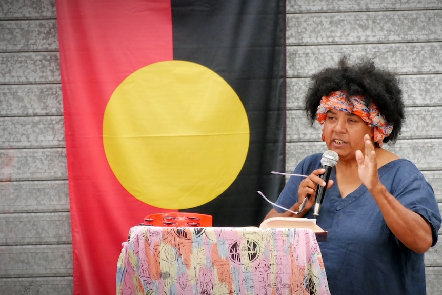 Woman with microphone and lectern in front of Aboriginal flag.