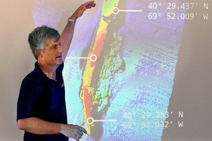A man with a black t shirt and grey hair is pictured pointing at a projected image of the wreckage of an ocean liner.