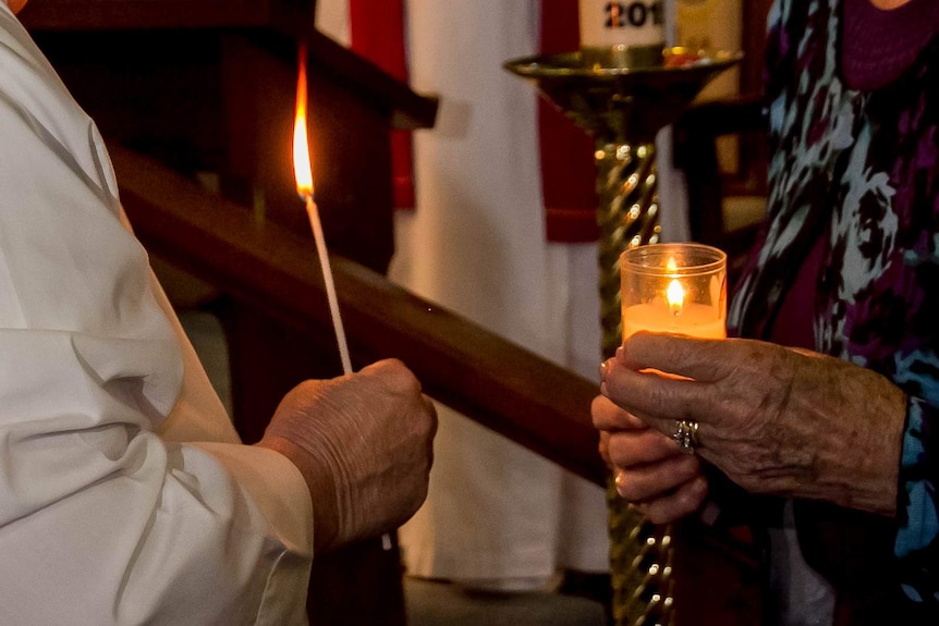 The hands of two women, one holding a lighted taper the other a lit candle inside a church