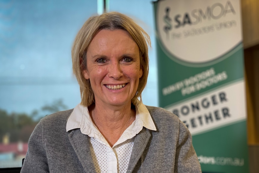 A woman with short blonde hair wearing a grey blazer standing in front of a sign for SASMOA