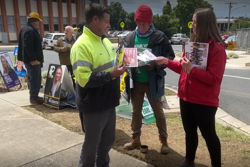 A man in a highly visible jacket takes a voting card from a young woman in a red blouse.  A hospital can be seen in the background