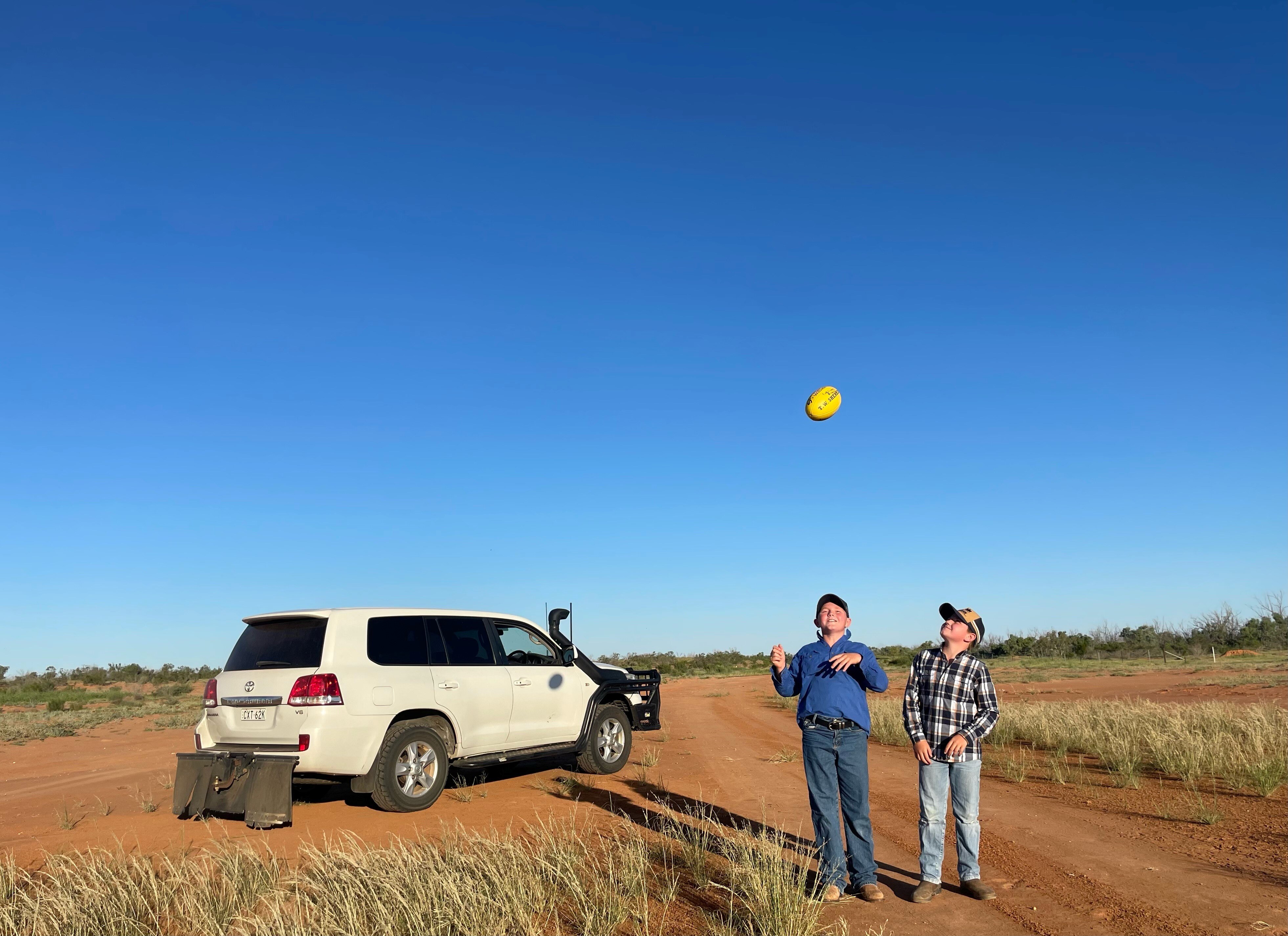 two young boys throwing a football in the air standing on a dirt track next to a car
