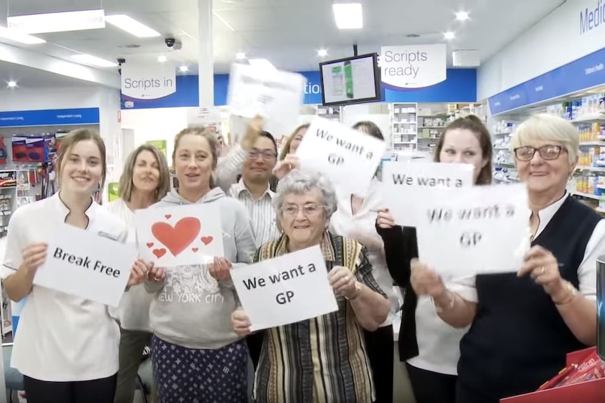 A group of people stand in a pharmacy and hold up signs saying 'We want a GP'.