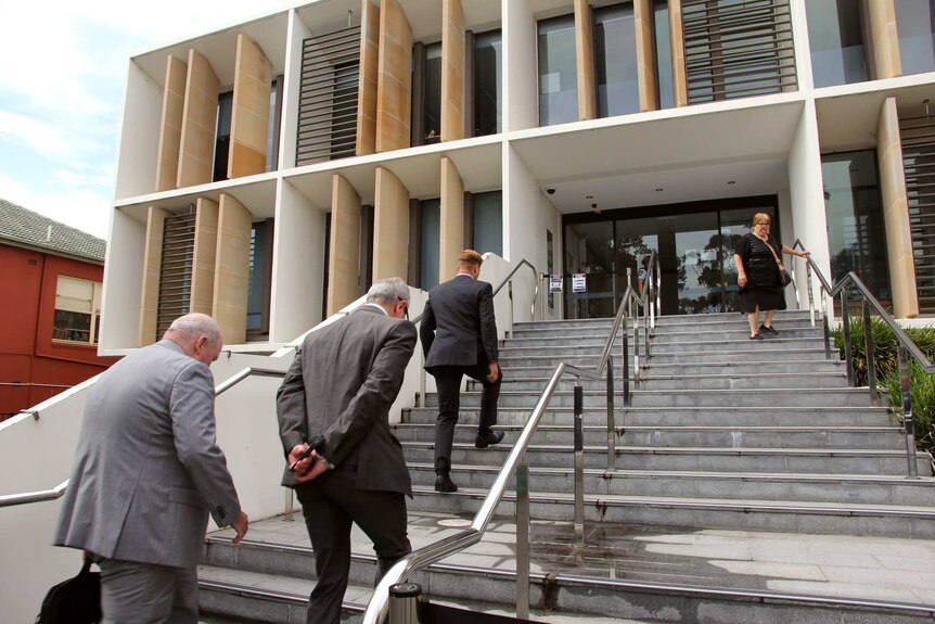 Three men walk up a flight of stairs towards a court building.