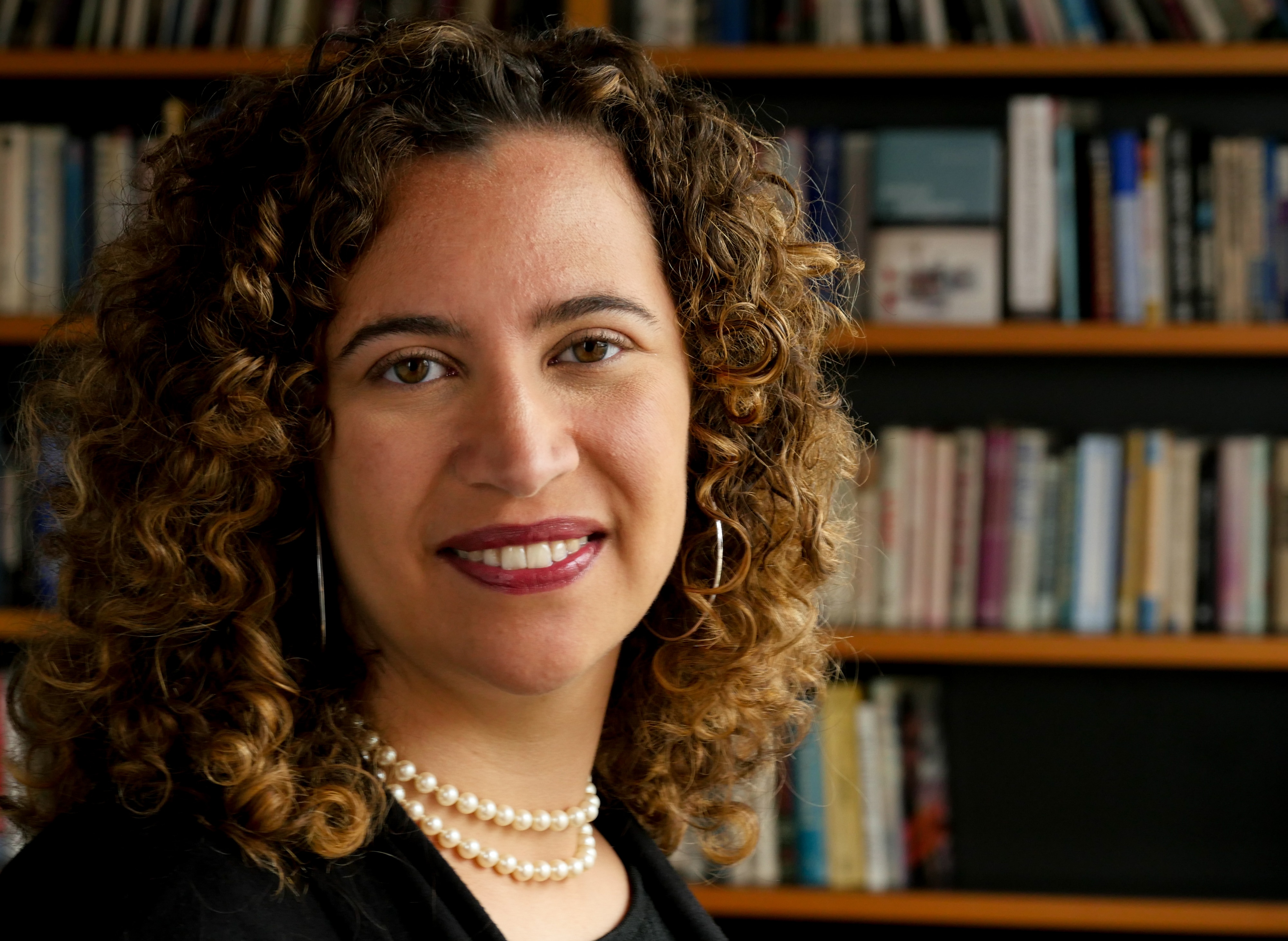 A woman with curly hair and hoop earrings stands in front of a bookshelf smiling.