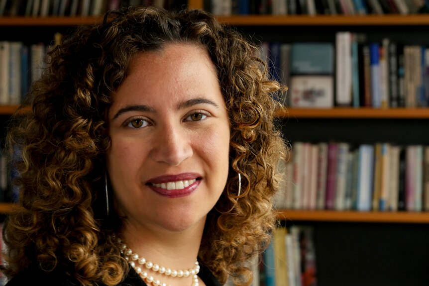 A woman with curly hair and hoop earrings stands in front of a bookshelf smiling.