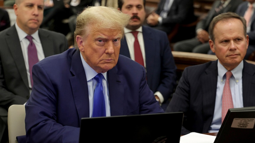 Donald trump frowns at a desk in a crowded courtroom 