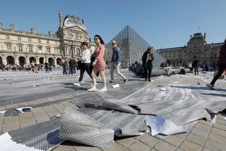 People walk over and look down at shredded sheets of grey and white dotted paper laid across paving stones in a grand courtyard.