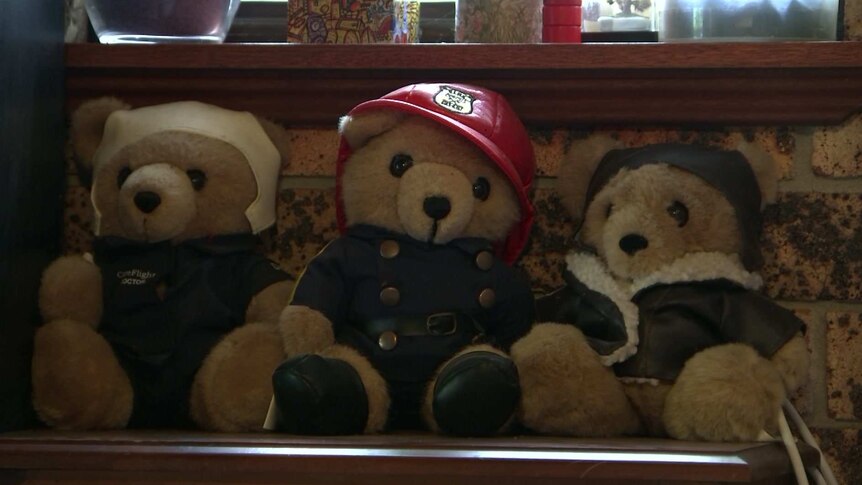 three teddy bears on a shelf in a row, the middle is dressed as firefighter