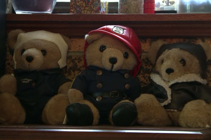 three teddy bears on a shelf in a row, the middle is dressed as firefighter