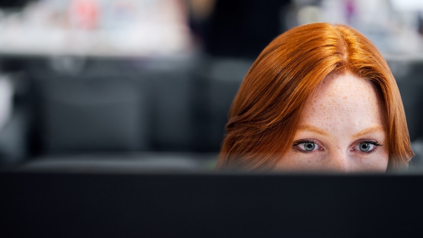 A young woman with red hair stares down at her computer, we only see the top half of her face.