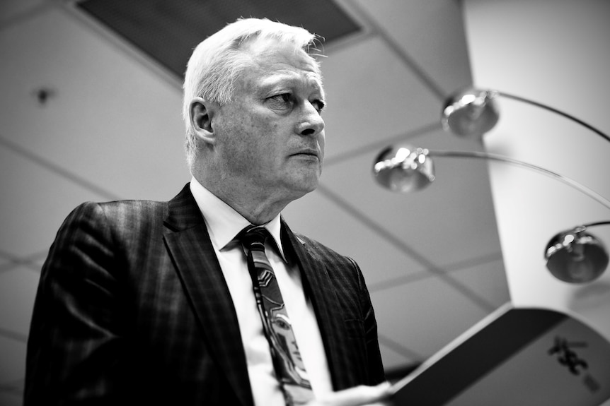 A side-on, black and white image of an older, silver-haired man looking straight ahead as he holds an open document