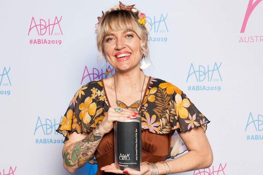 The author stands in front of a media wall holding her trophy, she is wearing butterfly clips and flowers in her hair.