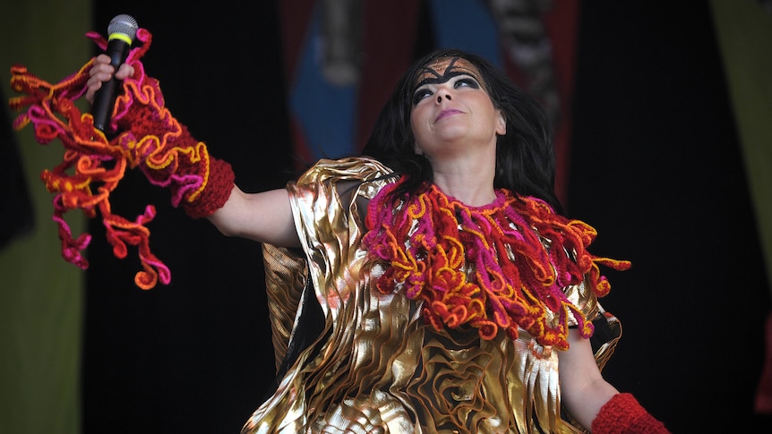 Bjork wears an intricate gold and orange costume and eye make-up. She holds up a microphone and looks up.