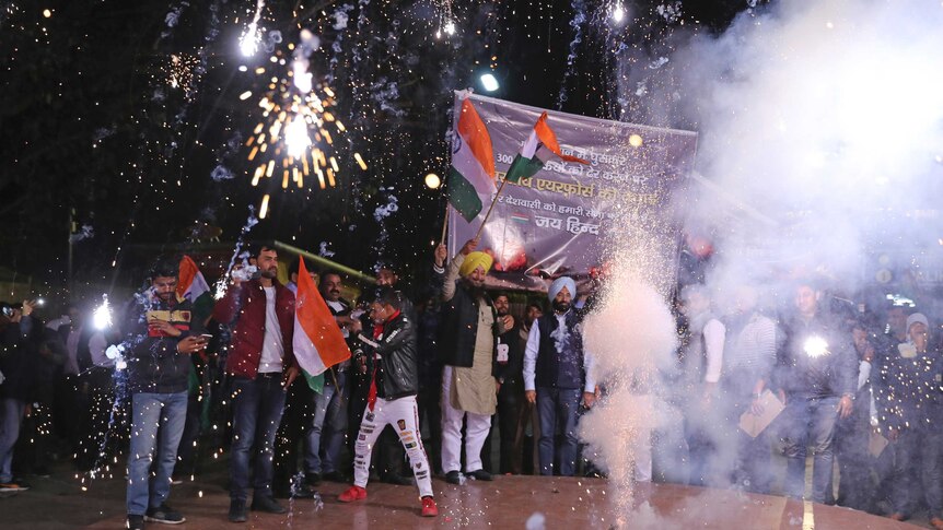 A group of people wave Indian flags and banners as a string of fireworks explode from a box on the ground.