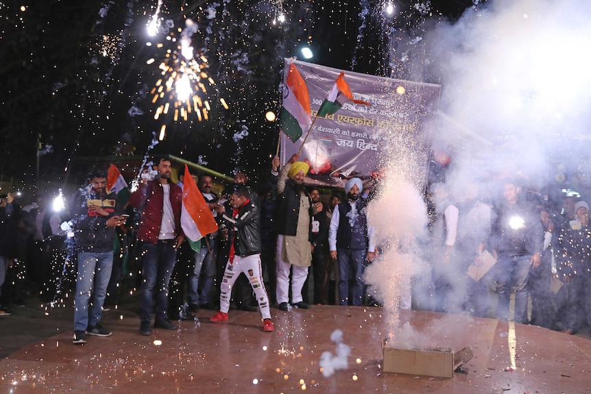 A group of people wave Indian flags and banners as a string of fireworks explode from a box on the ground.