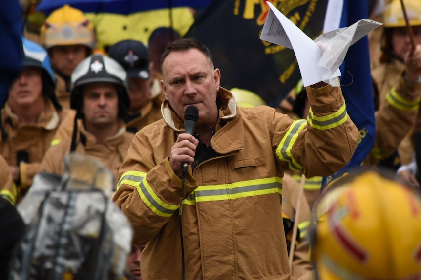 Peter Marshall in firefighting uniform with a microphone addresses a crowd of uniformed firefighters.