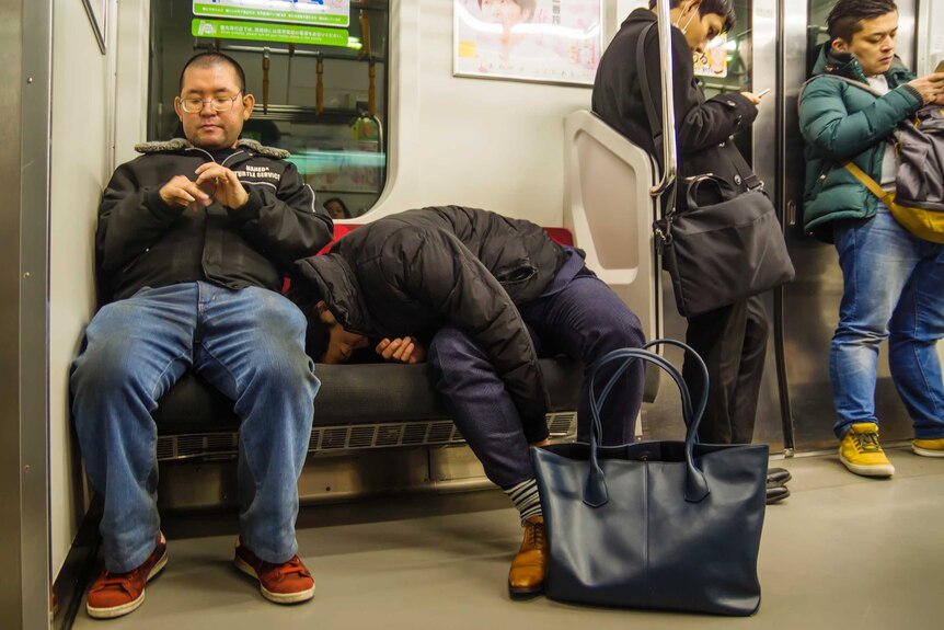 An office worker falls asleep in an unusual position on a public train late at night, as nonchalant commuters surround him