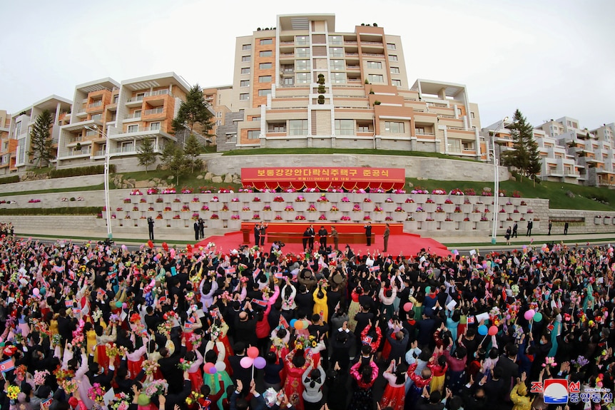 aerial view of people crowded outside an apartment building with a stage in front of it where a ribbon cutting ceremony is held
