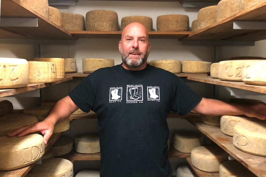 A person standing in their cellar (larder) with multiple cheese wheels all around