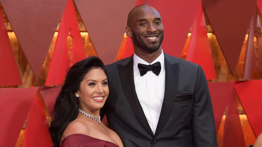 A short, smiling woman in a red dress stands next to a tall, bald, black man in a suit in front of a red Oscars background.