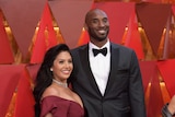 A short, smiling woman in a red dress stands next to a tall, bald, black man in a suit in front of a red Oscars background.