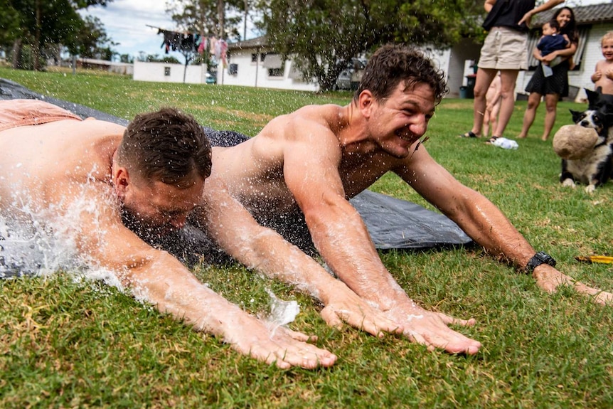 Two men in shorts slide along a wet black tarpaulin on grass.
