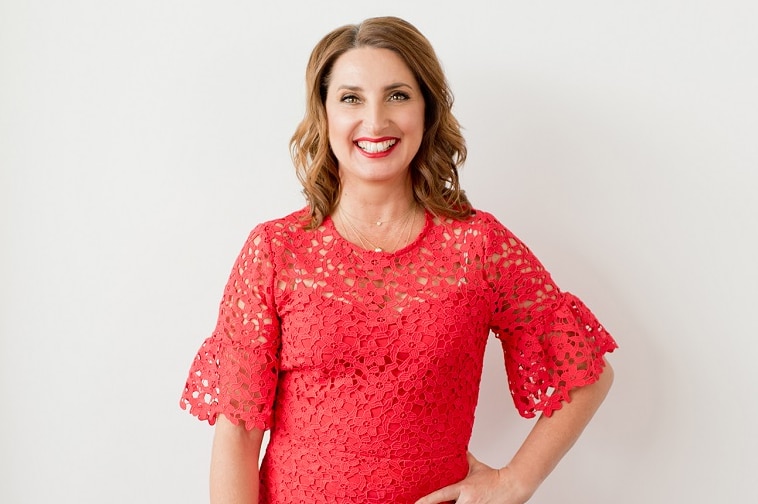 A woman with short curly brown hair in a coral pink dress standing against a white wall smiling with one hand on hip