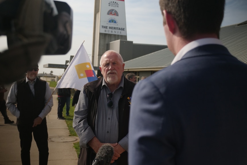 Bald man faces media to talk, union flags fly in background