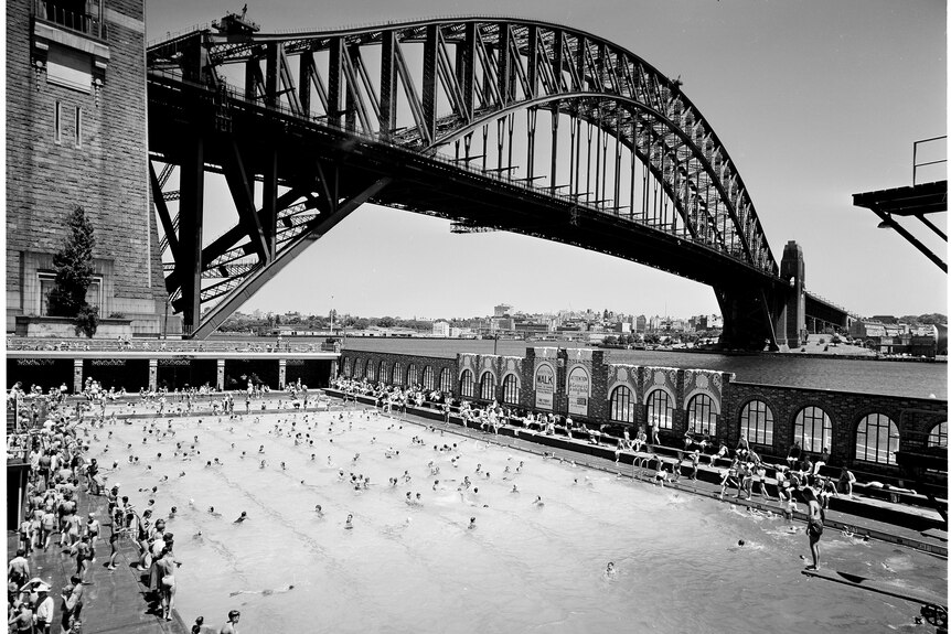 A black and white photograph of the North Sydney Olympic Pool next to the Sydney Harbour Bridge.