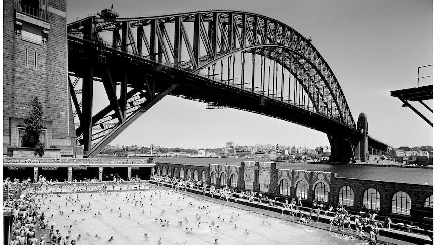 A black and white photograph of the North Sydney Olympic Pool next to the Sydney Harbour Bridge.