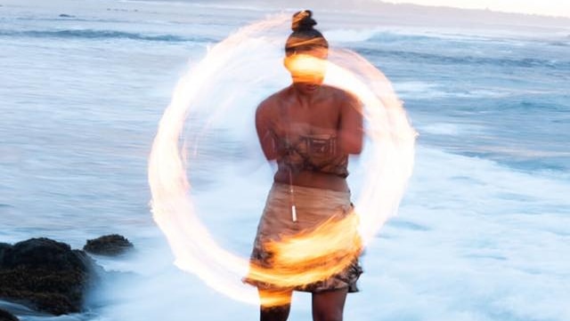 A long-exposure shot shows a ring of fire twirled by Moemoana Schwenke as she stands on a rock in front of the ocean.