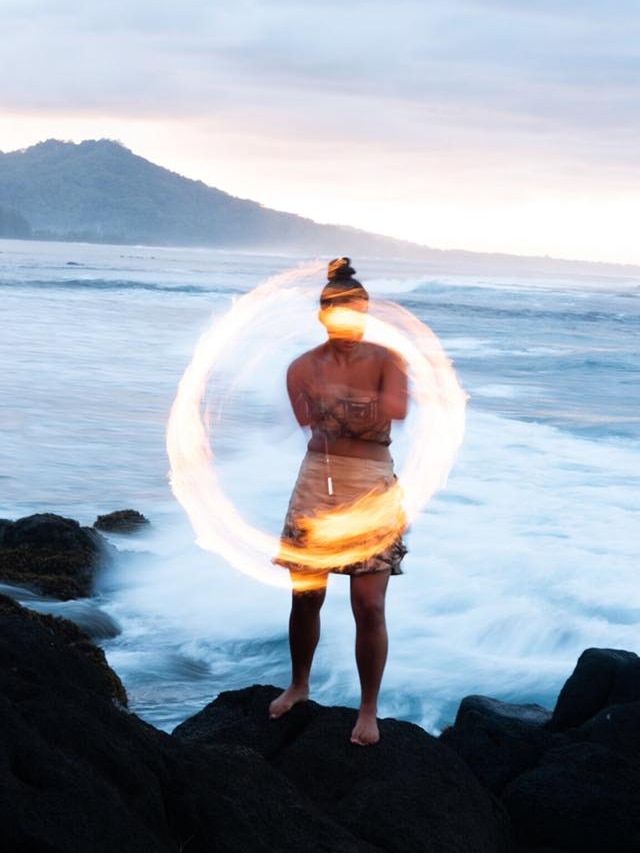A long-exposure shot shows a ring of fire twirled by Moemoana Schwenke as she stands on a rock in front of the ocean.