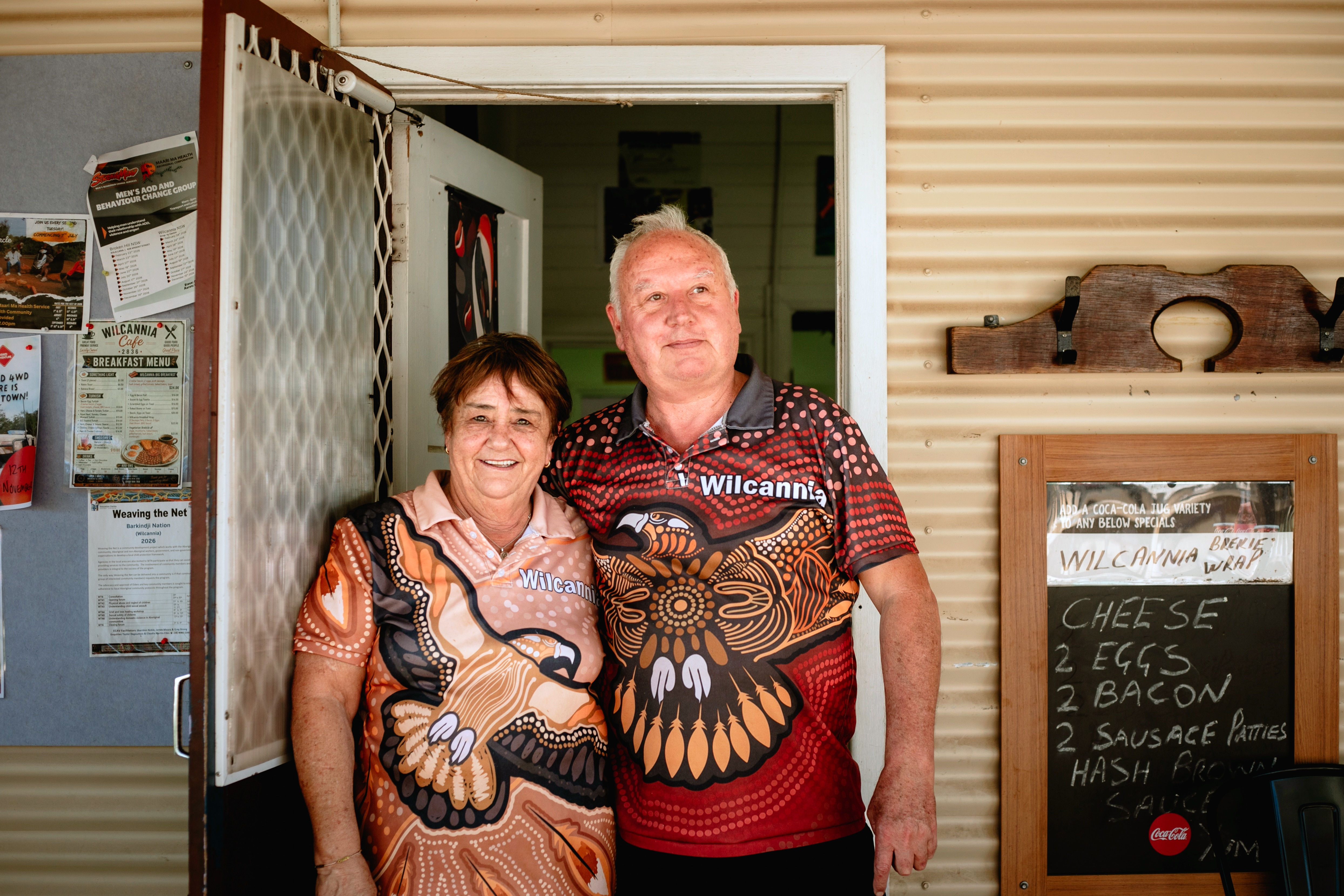 Shona and Conrad, an older couple, stand outside a cafe smiling.