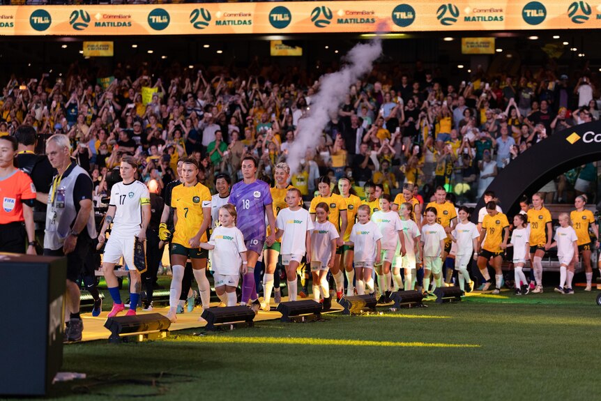 Two soccer teams, one wearing yellow and the other wearing white, walk onto the field with a big crowd behind them