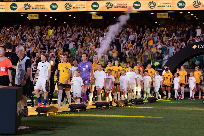 Two soccer teams, one wearing yellow and the other wearing white, walk onto the field with a big crowd behind them