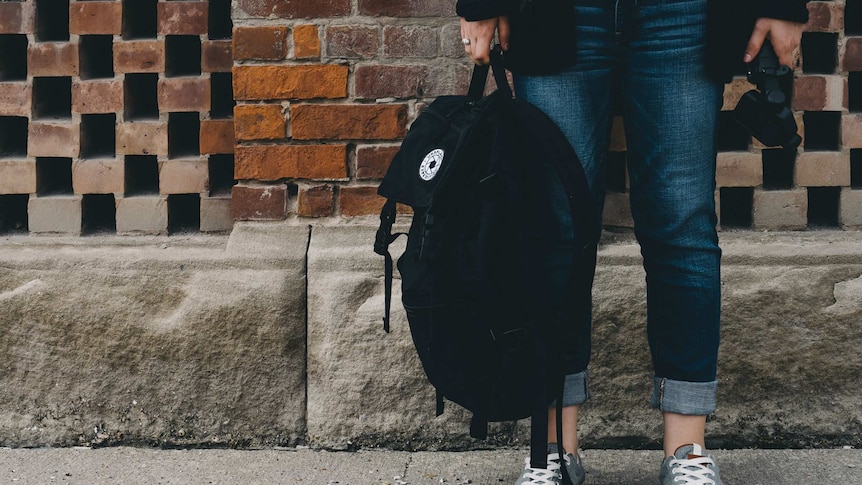 A teenager, standing against a wall, holds a backpack.
