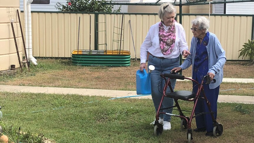 Gail Farrawell, carrying a blue watering can, walks beside her elderly mother, who is using a wheelie walker, in a backyard.