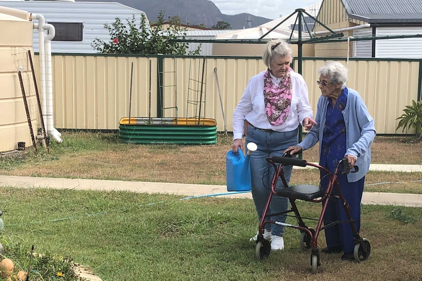 Gail Farrawell, carrying a blue watering can, walks beside her elderly mother, who is using a wheelie walker, in a backyard.