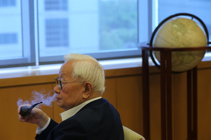 Man with white hair and glasses wearing a suit smokes a pipe in side profile.