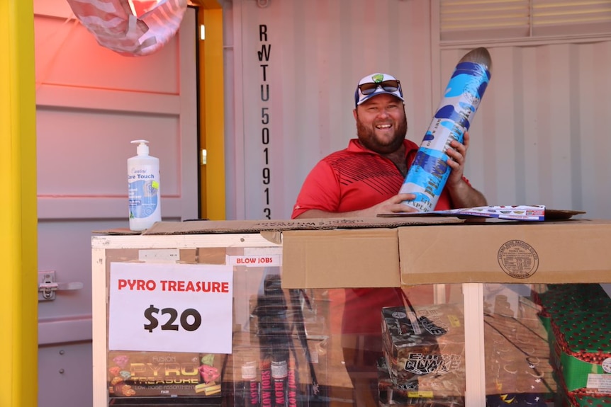 A smiling man in a baseball cap, standing behind a counter, holding a large bullet-shaped firework package.