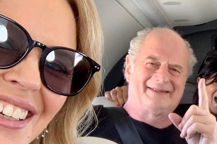 Blonde woman in dark sunglasses sits on a bus, greying, older man in black shirt behind her with index finger pointed