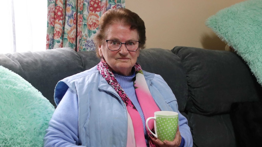 A woman in blue, with a pink scarf, holds a mug as she sits on a grey couch